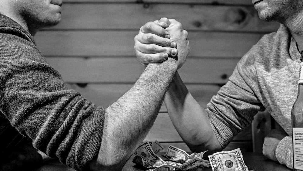 Two gentlemen arm wrestling on a table covered with money, signifying that if you do things correctly in the first place, you won't need to struggle.