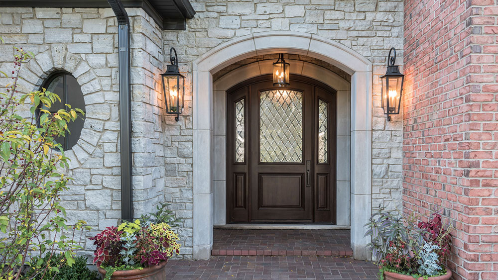 Arch top, single entry wood door with diamond divided lites and coordinating sidelites set into the covered exterior of a stone and brick home