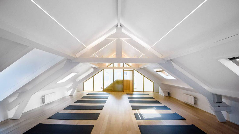 Yoga mats set up in 2 rows on a light wood floor in a long room with a vaulted ceiling and a wall of windows at the head of the room.