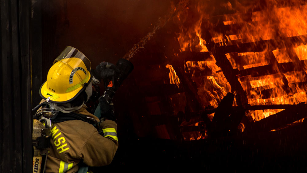 Firefighter with fire hose spraying a burning pile of wood pallets, signifying their exposure to harmful chemicals during training exercises