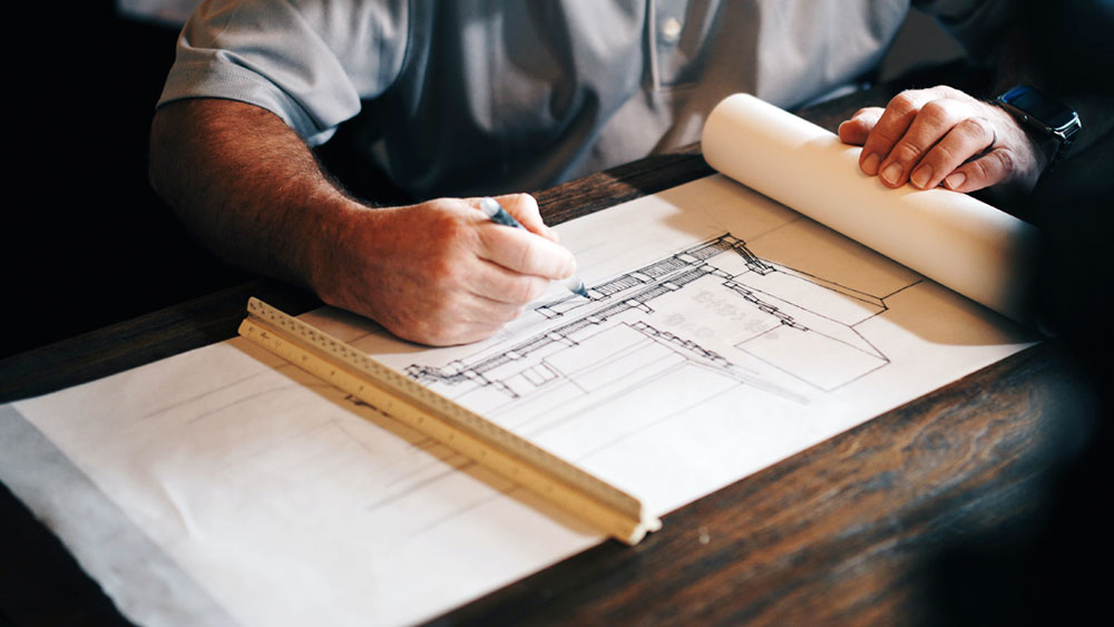 Architect in a gray shirt hand drawing building plans for a railing and staircase on a roll of paper with a ruler