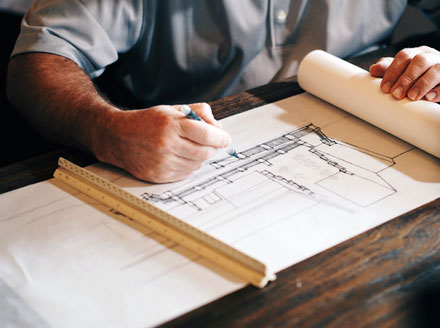 Architect in a gray shirt hand drawing building plans for a railing and staircase on a roll of paper with a ruler