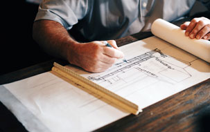 Architect in a gray shirt hand drawing building plans for a railing and staircase on a roll of paper with a ruler