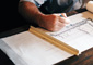 Architect in a gray shirt hand drawing building plans for a railing and staircase on a roll of paper with a ruler