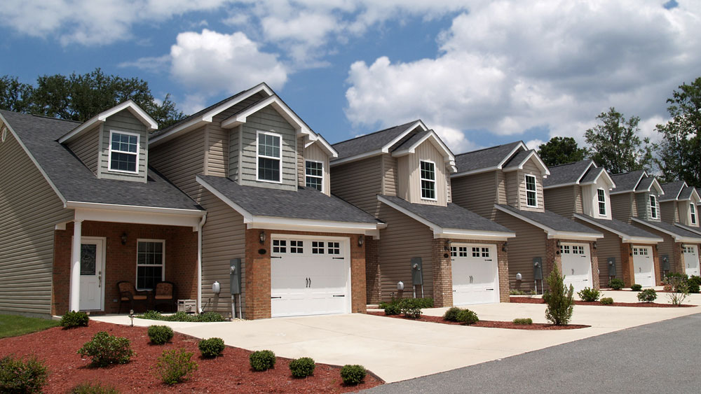 A row of suburban tract houses with identical architecture windows, brick facade, and siding along a sunny street.