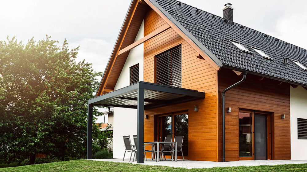 A modern passive house with slatted sun shades, black solar roof tiles, wood siding, and a metal overhang to shade the sliding glass door.