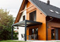 A modern passive house with slatted sun shades, black solar roof tiles, wood siding, and a metal overhang to shade the sliding glass door.