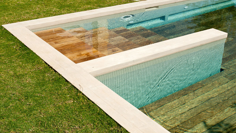 Moveable floor teak pool cover in the open position, revealing pool steps and adjustable depths of the blue tiled pool