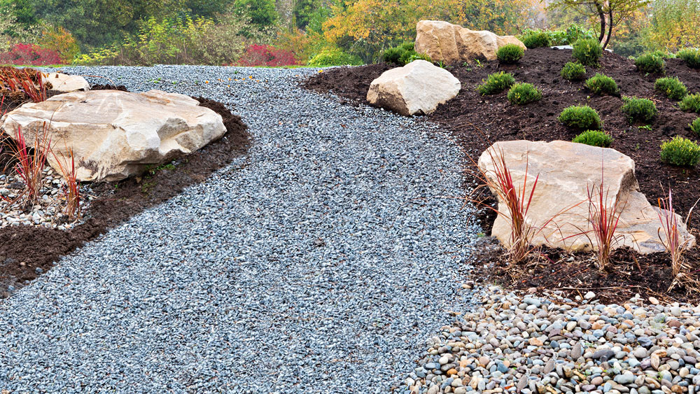 A xeriscape including a pathway of black basalt adjoined to a river rock drainage route, surrounded by dark bark mulched flowerbeds