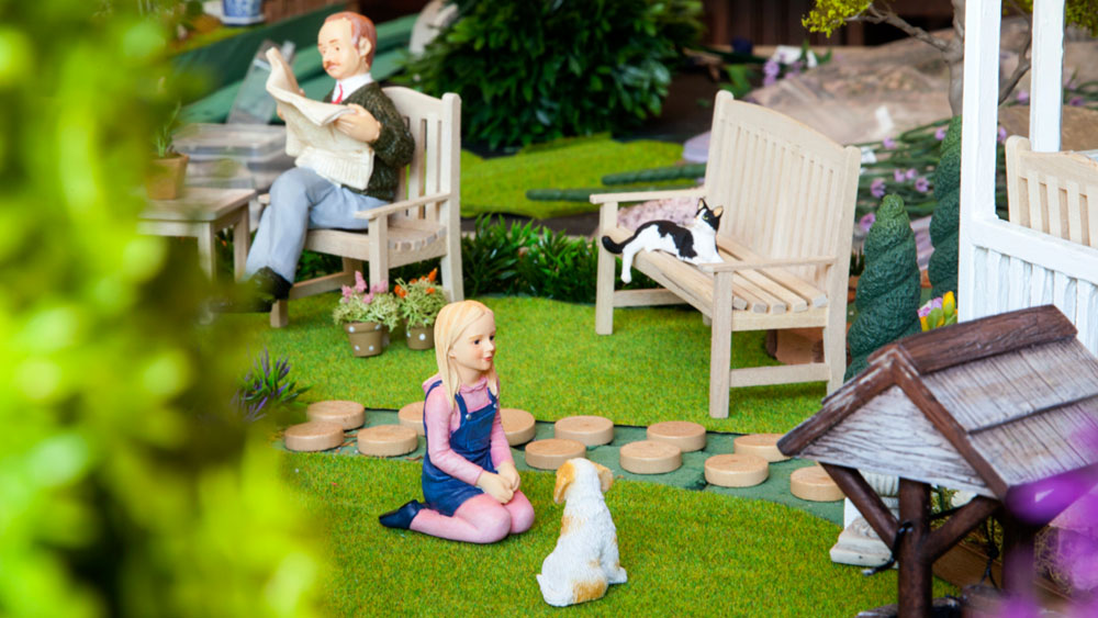 Dollhouse figurines of small girl kneeling in front of a dog and a man on a bench reading a newspaper in an outdoor courtyard