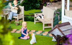 Turfgrass Oasis Dollhouse figurines of small girl kneeling in front of a dog and a man on a bench reading a newspaper in an outdoor courtyard