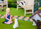 Dollhouse figurines of small girl kneeling in front of a dog and a man on a bench reading a newspaper in an outdoor courtyard