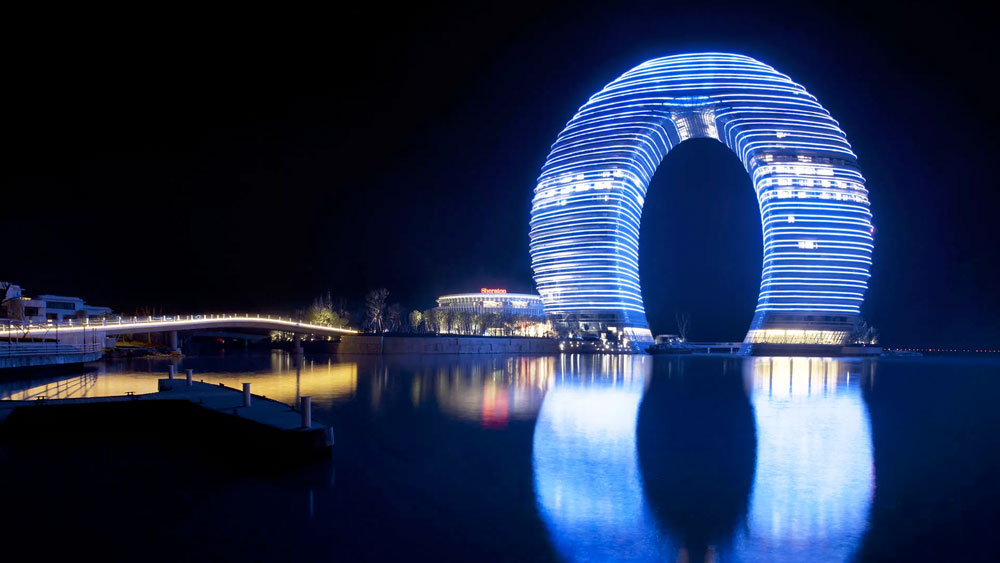 27-story, ring shaped Sheraton Hot Spring Resort in Huzhou, China lit blue at night with reflection on the water