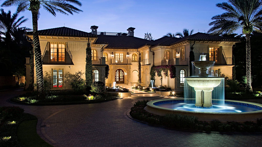 Evening exterior of stucco Mediterranean mansion in Naples, Florida with circular paver driveway surrounding a large fountain.