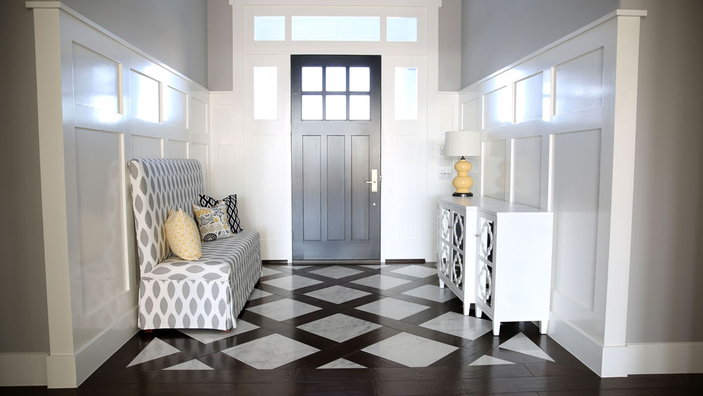 Bright traditional foyer with white paneled walls, black front door, and wide wood plank and marble flooring in a diamond pattern.