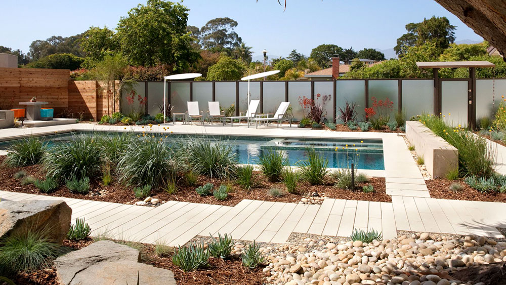 A modern, rectangular pool surrounded by a xeriscaped yard with ornamental grasses, river rocks, and wood mulch.